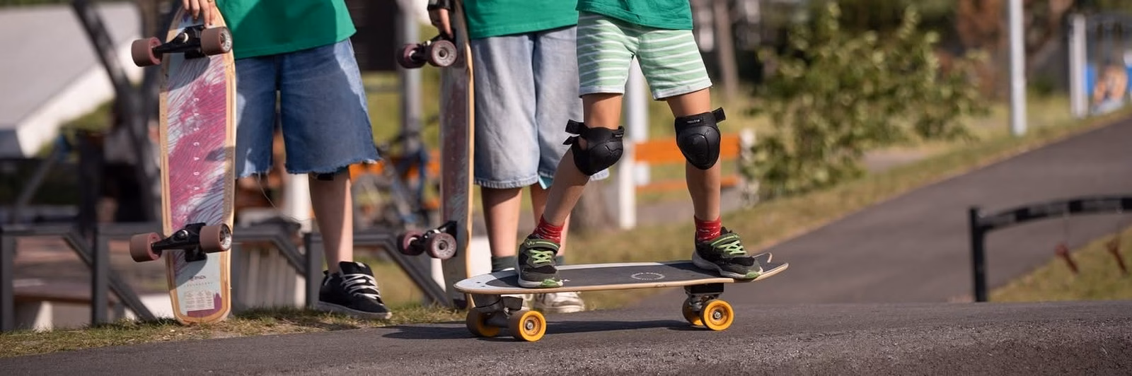 Troje dzieci z deskami surfskate na pumptracku, jedno jedzie, dwoje trzyma deski pionowo